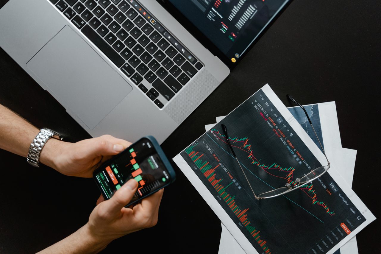 services-04 Hands using smartphone beside laptop with stock charts, showcasing digital trading.